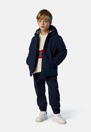 Giacca navy con cappuccio sopra un maglione crema con strisce rosse e blu, abbinata a pantaloni cargo scuri e scarpe da ginnastica bianche.