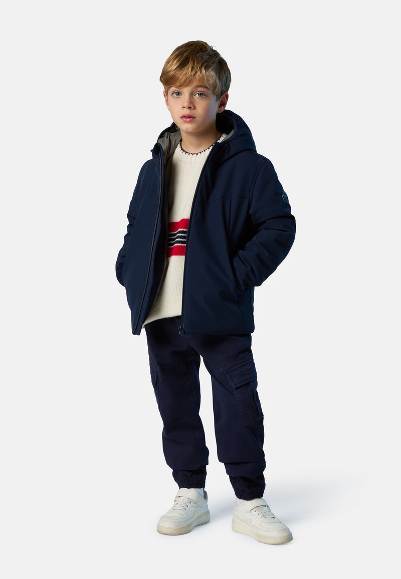 Veste marine à capuche portée sur un pull crème avec des rayures rouges et bleues, associée à un pantalon cargo foncé et des baskets blanches.