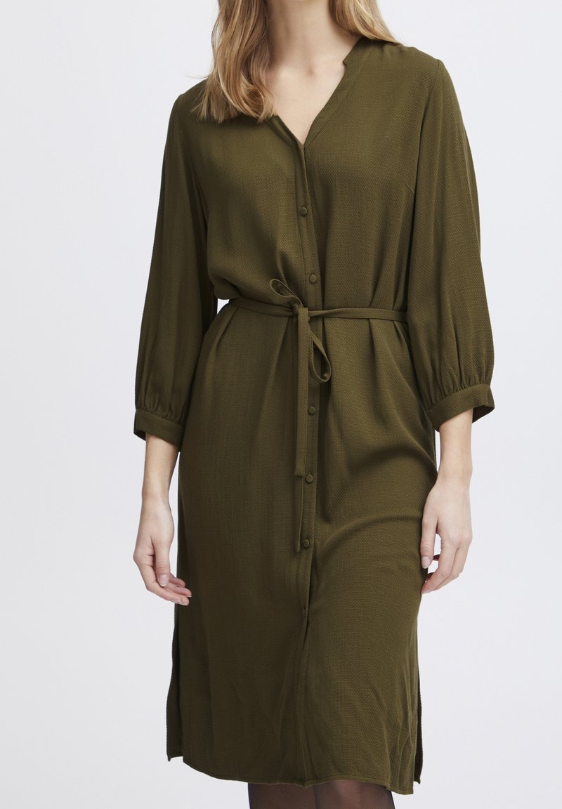 Femme portant une robe boutonnée à manches longues de couleur vert olive avec une ceinture en tissu nouée à la taille, montrée du cou jusqu'au milieu du mollet.