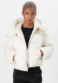 Pufferjack in off-white met een hoge capuchon en ritssluiting. Heeft een gevoerd ontwerp en twee voorkruiken, waardoor het een zachte textuur biedt.