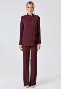 Blusa avvolgente bordeaux con fiocco laterale, abbinata a pantaloni a gamba larga coordinati. Tessuto liscio con maniche lunghe. Décolleté beige con punta.