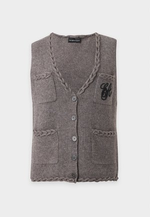 Kvinde iført grå strikket knapvest over sort top med korte ærmer, blå vide jeans og sorte spidse sko, stående mod hvid baggrund.