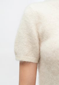 Pulover tricotat cu mâneci scurte, de culoare bej deschis, realizat dintr-un material moale și pufos, cu manșete ribanalate. Prezintă o croială lejeră și o textură fină.