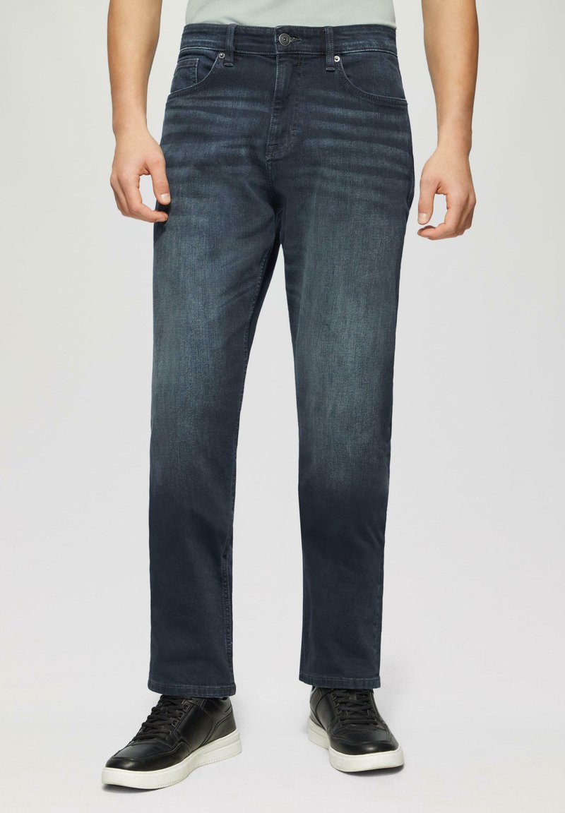 Jean en denim bleu foncé avec une coupe droite, présentant des poches avant, une fermeture à bouton et un léger effet délavé. Portés avec des baskets noires.