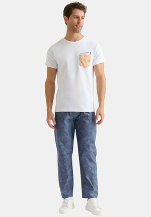 Maglietta bianca a maniche corte con taschino beige a motivo, abbinata a pantaloni scuri blu a motivo e scarpe da ginnastica bianche.