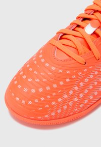 Pomarańczowy but piłkarski z teksturowaną, syntetyczną cholewką, białymi wzorzystymi akcentami i sznurowadłami. Okrągły nosek i amortyzowana podeszwa.