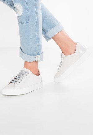 Persoon die lichtblauwe opgerolde jeans en lichtbeige suède sneakers met grijze elastische bandjes draagt, zittend tegen een witte achtergrond.