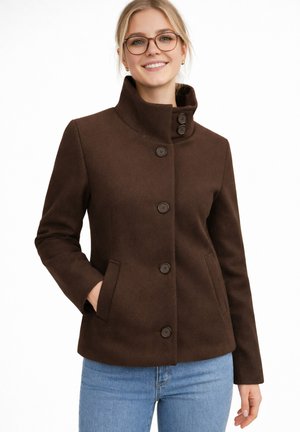 Młoda kobieta nosząca okulary i brązowy płaszcz na guziki z wysokim kołnierzem, pozująca z jedną ręką w kieszeni i uśmiechająca się.