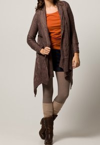 Brązowy dziergany kardigan o luźnym kroju z warkoczowym wzorem, zestawiony z pomarańczową bluzką, ciemnymi szortami i brązowymi botkami sięgającymi do kolan.