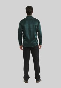 Chemise longue en satin vert à col classique, avec des poignets boutonnés et une texture lisse, assortie à un pantalon noir et des chaussures.
