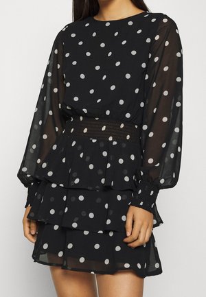 Zwarte jurk met witte polkadots, doorschijnende lange mouwen, elastische tailleband en gelaagde rok, gedragen door een persoon van de nek tot halverwege de dij.