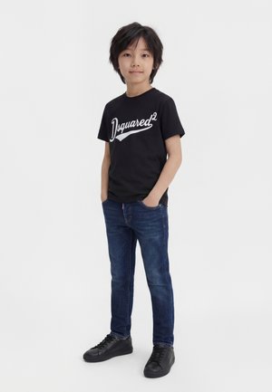 Dreng stående med hænderne i lommerne iført sort "Dsquared²" t-shirt, mørkeblå jeans og sorte sneakers på hvid baggrund.