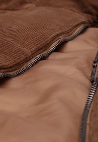 Gros plan d'une veste en velours côtelé marron avec une fermeture éclair noir et blanc partiellement ouverte, montrant une doublure intérieure lisse.