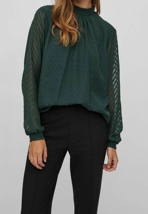Vrouw draagt een donkergroene transparante blouse met lange mouwen en een getextureerd chevronpatroon en zwarte hoge taillebroek.