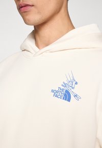 Dukserica krem boje s prednjim džepom kangura. Na lijevoj strani prsa nalazi se plavi logo i grafika skijaša. Mekani materijal, kapuljača s podesivim remenjem.