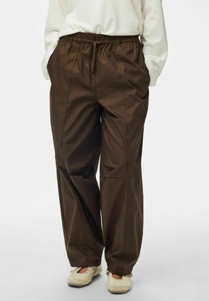 Personne portant un pantalon marron ample avec taille élastique et cordon, un haut blanc à manches longues et des chaussures plates de couleur claire.