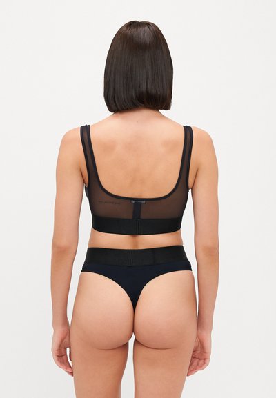Černá průhledná podprsenka s širokými popruhy a odpovídajícími černými tangy. Hladký materiál, minimální kovové prvky a elastický pásek v pase.