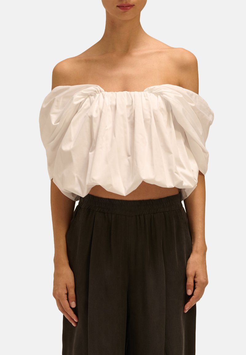 Biała bluzka z odkrytymi ramionami, z marszczonym materiałem i skróconym krojem, w połączeniu z luźnymi, ciemnoszarymi spodniami z elastycznym paskiem.