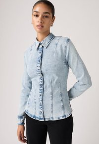 Γυναίκα που φοράει εφαρμοστό, ανοιχτό μπλε πουκάμισο από acid-wash τζιν με κουμπιά μπροστά και μαύρο παντελόνι, στέκεται με το ένα χέρι λυγισμένο.