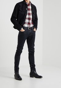 Zwarte denimjas over een rood-witte geruite shirt, gecombineerd met donkere jeans en zwarte leren enkellaarsjes. Een eenvoudige riem maakt de outfit af.