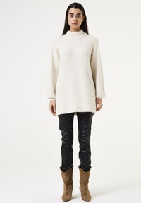 Lange, crèmekleurige fuzzy gebreide trui met een hoge hals, gecombineerd met glanzende zwarte broek en bruine lange laarzen met bloemenborduursel.