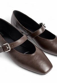 Zapatos Mary Jane de cuero marrón con hebillas plateadas y puntas redondeadas en forma de cuadrado, exhibidos sobre una superficie blanca.