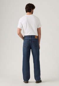 Jeans de mezclilla azules para hombre con corte recto, que cuentan con bolsillos traseros, un parche de cuero en la cintura y una textura suave. Combinados con una camiseta blanca.