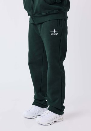 Personne portant un pantalon de survêtement vert foncé avec le logo blanc PXP et des chaussures de sport blanches, debout devant un fond uni.