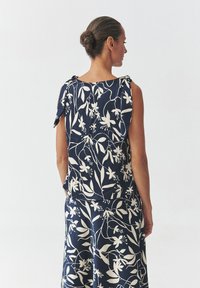 Rochie navy cu un model floral alb mare, material elastic, fără mâneci și cu detalii de legare pe umeri. Potrivită pentru ținute casual.