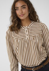 Donna con capelli ricci che indossa una camicia a righe beige e bianca con bottoni, jeans scuri e una cintura marrone, su uno sfondo neutro.
