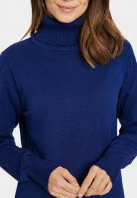 Maglione blu a collo alto in tessuto a costine con maniche lunghe e polsini aderenti, caratterizzato da un alto colletto ripiegato.