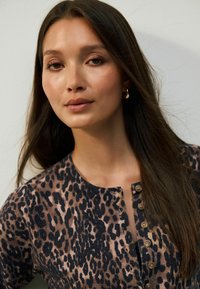 Jeune femme avec de longs cheveux bruns portant des boucles d'oreilles en or et un haut à boutons en imprimé léopard, légèrement tournée vers la gauche sur un fond uni.