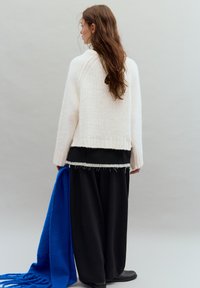 Weißer Strickpullover, schwarzes Schicht-Top, lockere schwarze Hose, blauer Schal über den Arm gelegt, glatte Textur und entspannte Passform.