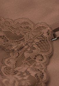 Bruine stof met complexe kantdetails langs de rand. Het kant heeft bloemmotieven en delicate texturen, wat de zachtheid van het materiaal versterkt.