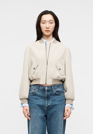 Młoda kobieta ubrana w beżową kurtkę bomberkę na niebieską koszulę i wysokowalne niebieskie dżinsy, stojąca na tle jasnego, jednolitego tła.