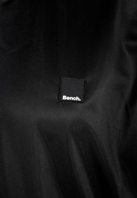 Černá textilní taška s obdélníkovou černou nálepkou s nápisem "Bench." v bílém písmu. Materiál je hladký s jemnou texturou.