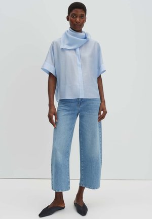 Vrouw die een lichtblauw loszittend shirt met korte mouwen met een sjaaldetail draagt, cropped jeans met wijde pijpen en zwarte puntige platte schoenen, staand tegen een witte muur.