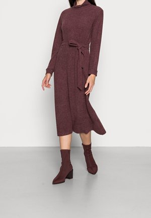 Bordeauxrode gebreide jurk met lange mouwen, hoge hals en een ceintuur in de taille. Draagt bijpassende enkellaarsjes met hak, met een gladde textuur.