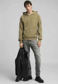 Felpa con cappuccio verde oliva con tasca frontale, polsini e orlo a costine. Jeans grigi e giacca di pelle nera tenuta nella mano sinistra. Stivali neri completano il look.