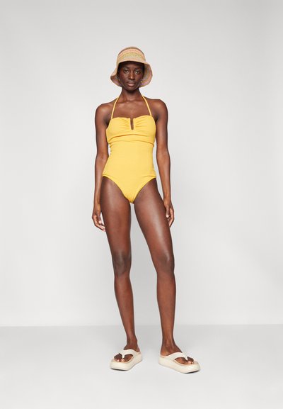 Maillot de bain une pièce jaune avec un corsage froncé et un détail en œillet, associé à un chapeau seau beige et des sandales. Texture lisse, design ajusté.
