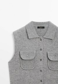Gilet sans manches en tricot gris avec col replié, fermeture à boutons sur le devant et deux poches poitrine boutonnées sur fond blanc.