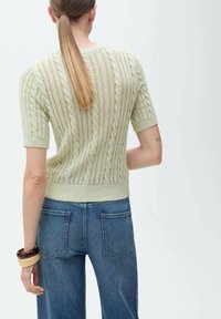 Pulover scurt din tricot, de culoare verde deschis, cu mâneci scurte, prezentând un model texturat și tiv ribotat, asortat cu blugi din denim albastru cu talie înaltă.