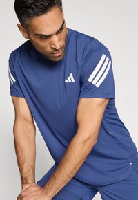 Camiseta deportiva azul de manga corta, con detalles de tres franjas blancas en los hombros y un pequeño logotipo en el pecho. Textura suave.