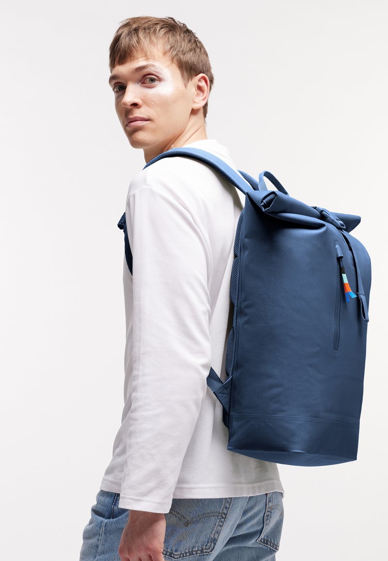 Junger Mann mit weißem langärmligem Hemd und Jeans, der einen großen blauen Rucksack trägt und über die Schulter gegen einen einfarbigen Hintergrund schaut.