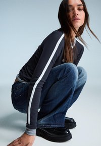 Hosszú ujjú sötétkék ing fehér oldalcsíkokkal, kék bő szárú farmer, és fekete platform cipők. A modell guggoló pózban, világos háttér előtt.