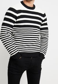 Pull en maille à rayures noir et blanc avec un col rond à côtes et des poignets côtelés, présentant une texture épaisse et des motifs horizontaux.