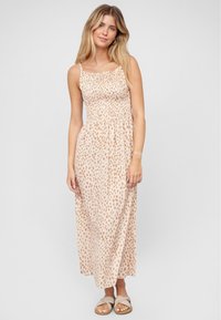 Jeune femme debout, portant une robe maxi sans manches beige avec des taches brunes et des sandales plates beiges, sur un fond uni clair.