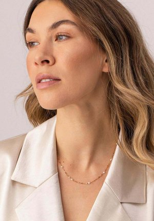 Frau mit welligem hellbraunem Haar, die eine Perlenkette und eine goldene Gliederkette sowie ein cremefarbenes Satinblazer trägt und nach links schaut.