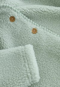 Fuzzy mintgrøn fleece-stof med en tekstureret overflade, rundede detaljer og to træknapper til lukning.
