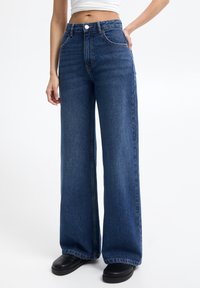 Jean en denim bleu à jambes larges avec une taille haute, des coutures contrastantes, un design à cinq poches et une finition soignée. Associé à des chaussures noires.